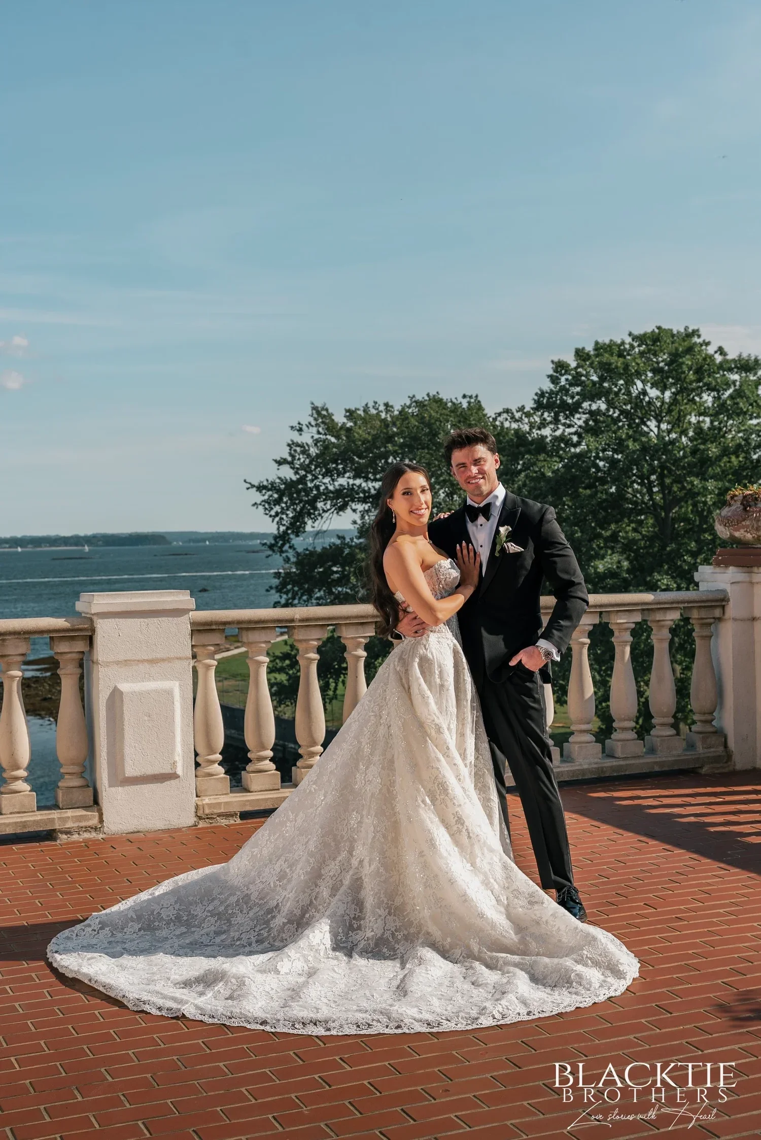 Leah and Nicholas standing close on the terrace, her lace train fanned across the brick, the white balustrade, trees and the Sound behind them under a blue sky
