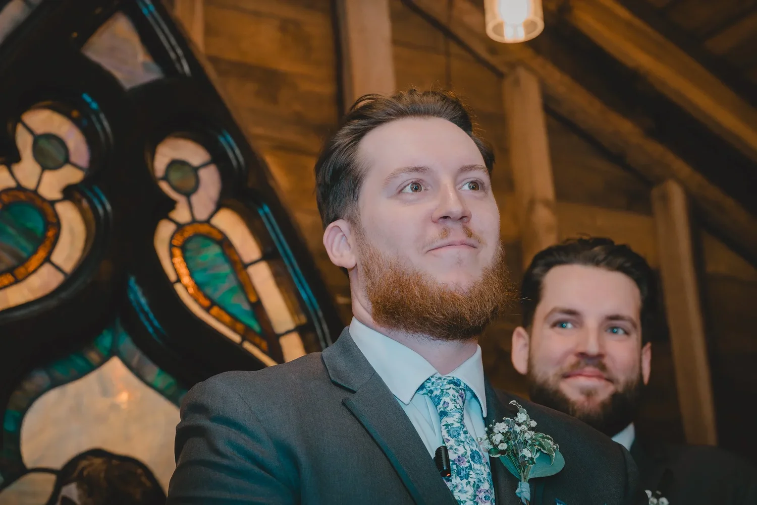 Christian smiling at the front of the Loft Chapel beside the stained glass window, with a groomsman behind him