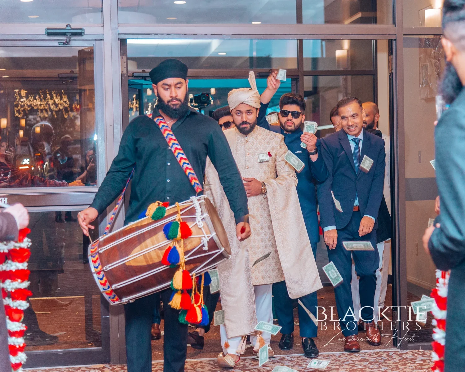 A dhol player leading the groom and his party in through the glass entrance doors of the hotel at night while dollar bills fall through the air
