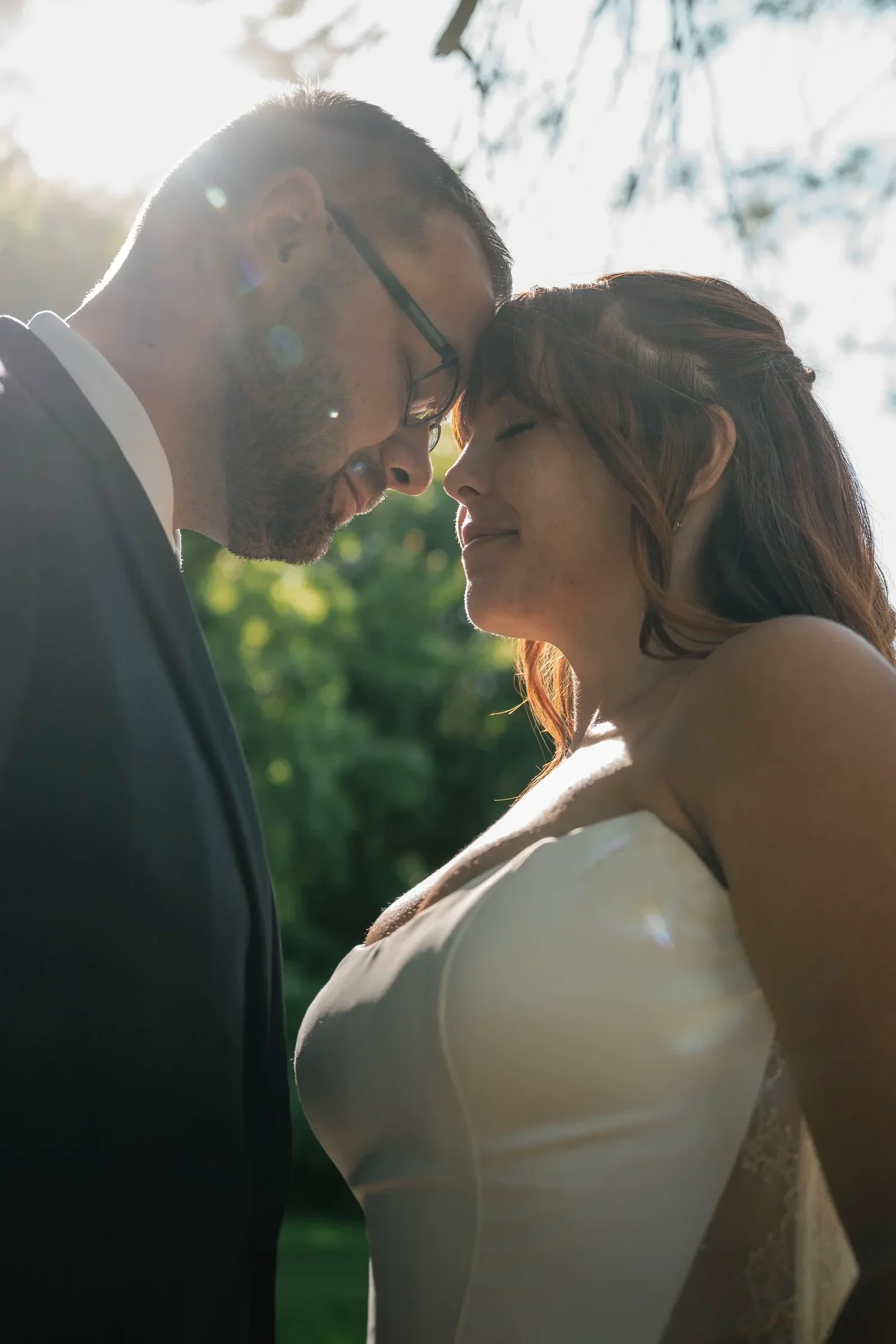 A close portrait of Danielle and Zach forehead to forehead, backlit by late afternoon sun through the trees at Traditions at the Glen