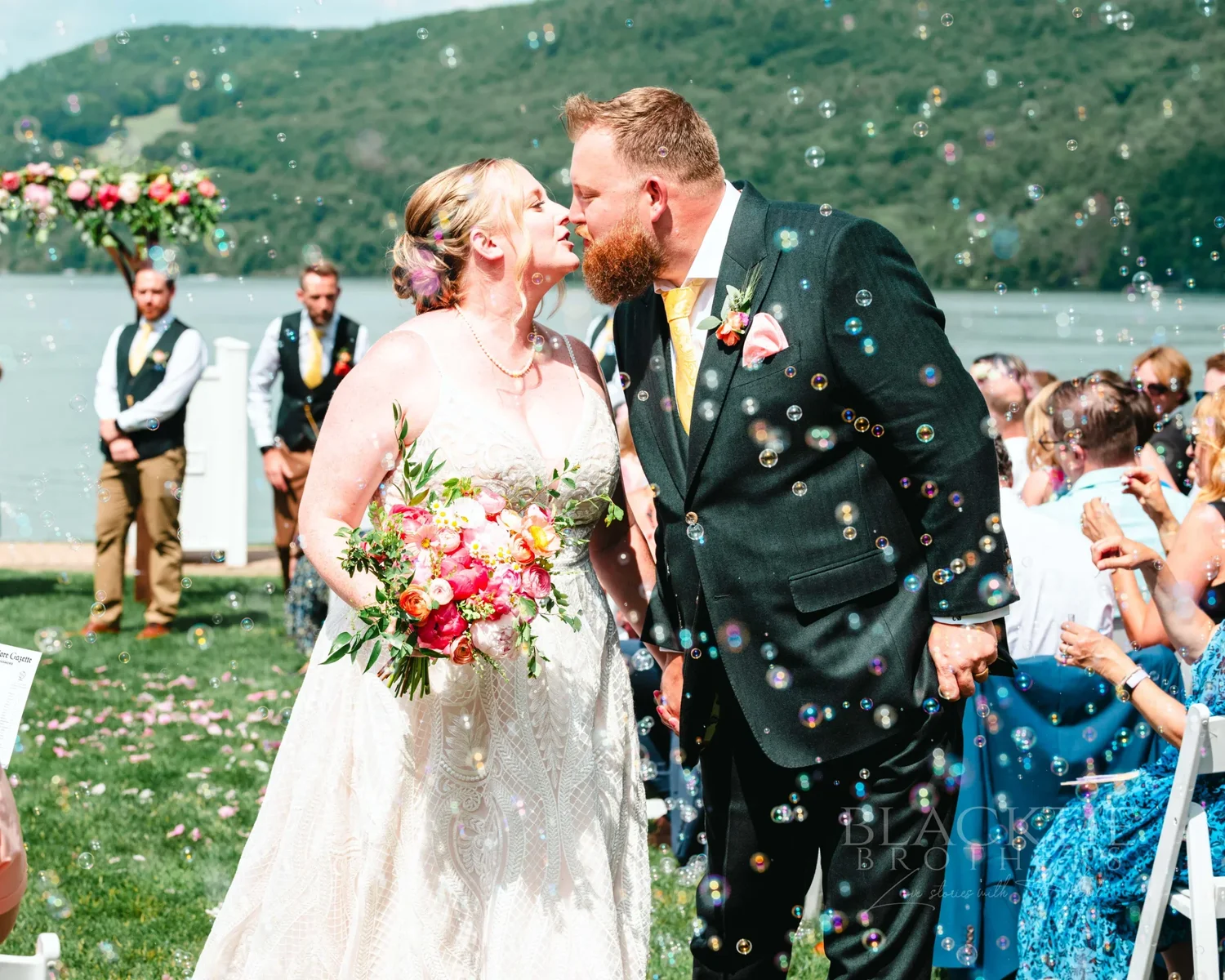Amanda and Chris kissing as guests throw confetti after their lakeside ceremony at The Otesaga