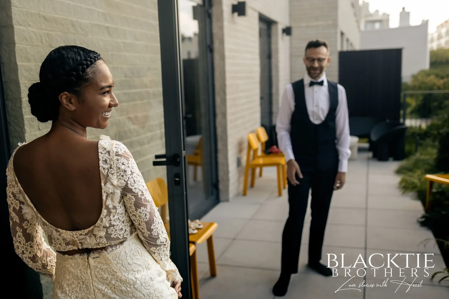 Ciara smiling back over her shoulder during the first look on the terrace, Drew grinning a few steps away beside yellow chairs
