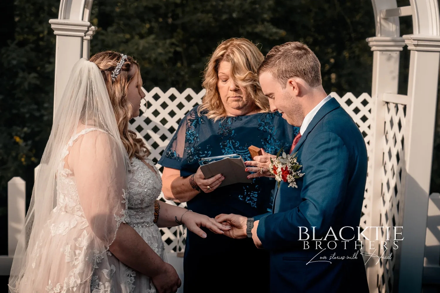 Jared placing the ring on Jennifer's finger at the arbor while the officiant, in a navy dress, holds the ceremony book