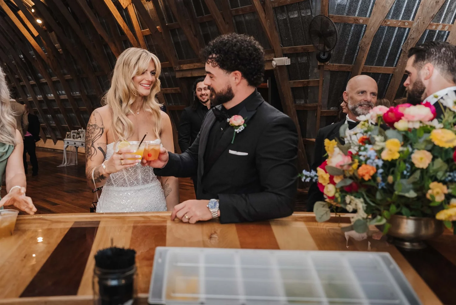 Kari and Josh smile at each other and touch glasses at the wooden barn bar.