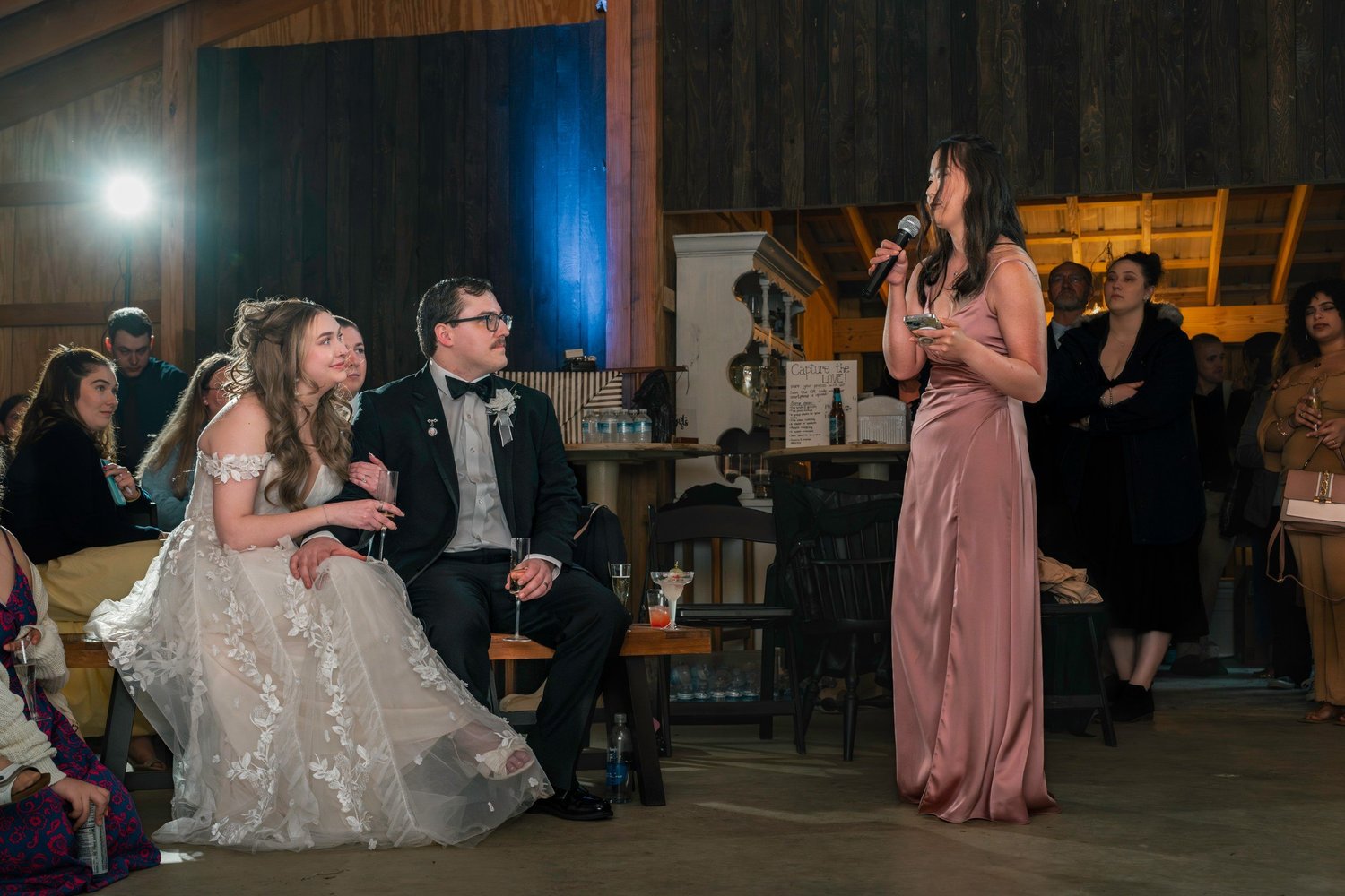 A bridesmaid in a mauve dress giving a toast while Lindsey and Noah listen from their seats