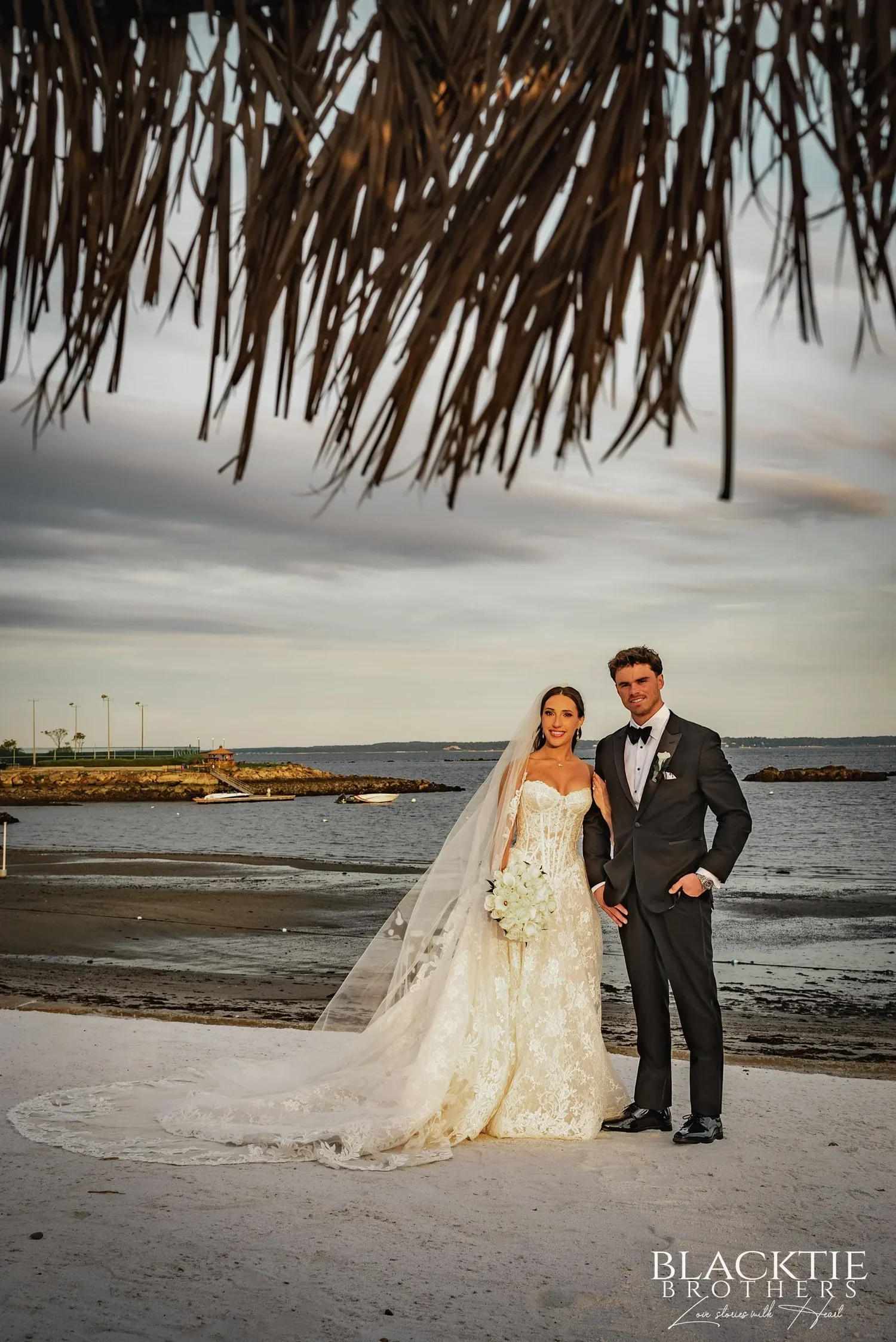 Leah and Nicholas standing on the beach at dusk, her veil and train across the sand, rocky outcrops and calm water behind them, framed by the thatched fringe of a beach umbrella