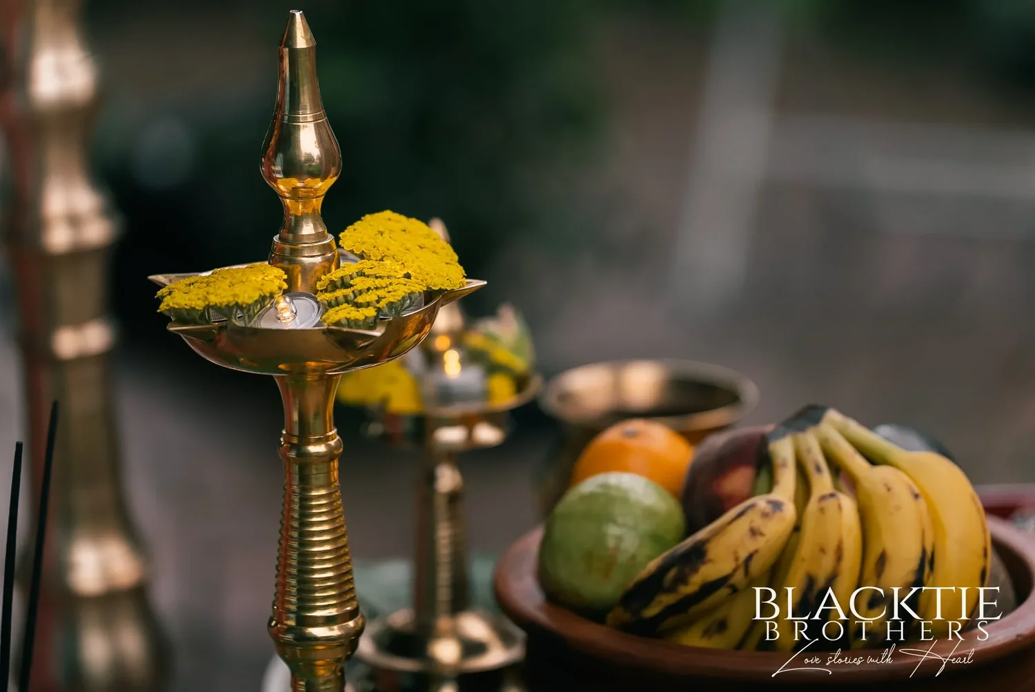 A tall brass oil lamp topped with yellow flowers beside a bowl of bananas, oranges and apples at the ceremony