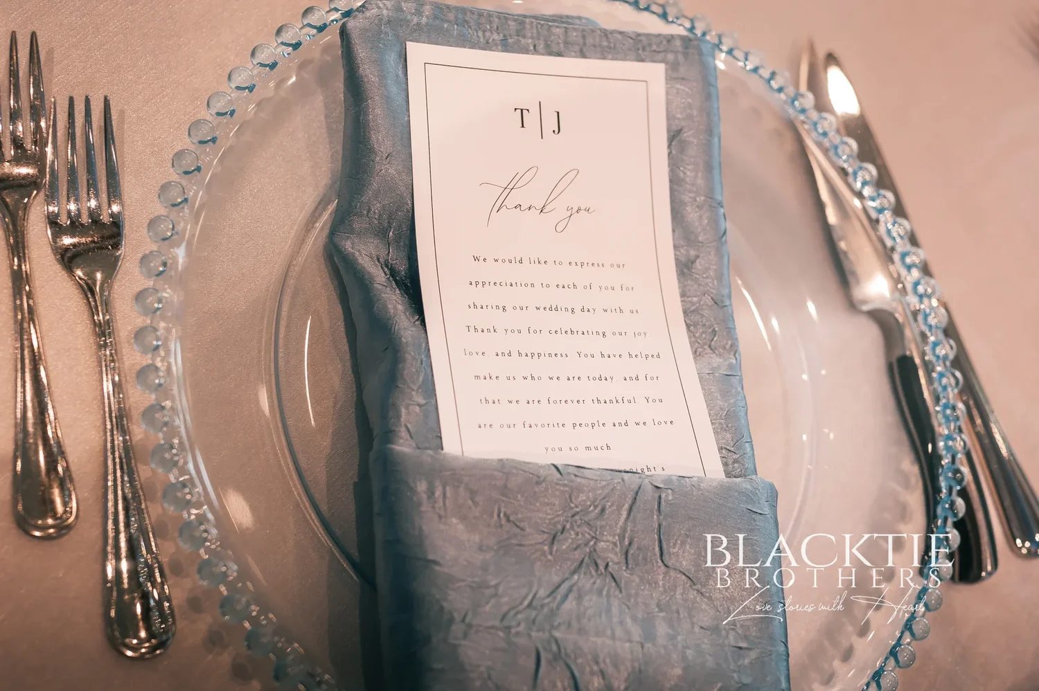 A place setting at Taylor and Jake's reception: a glass charger with a beaded rim, a dusty blue napkin and a printed thank-you card with the letters T and J