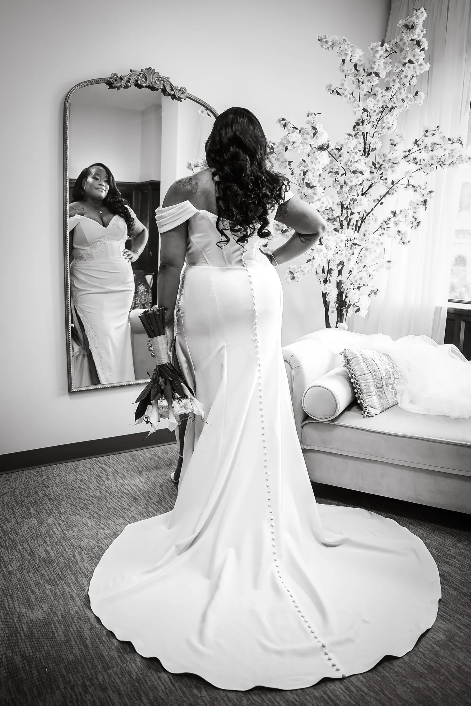 Black and white photograph of Tiffany from behind in her fitted gown and long train, her reflection smiling back from an ornate mirror.