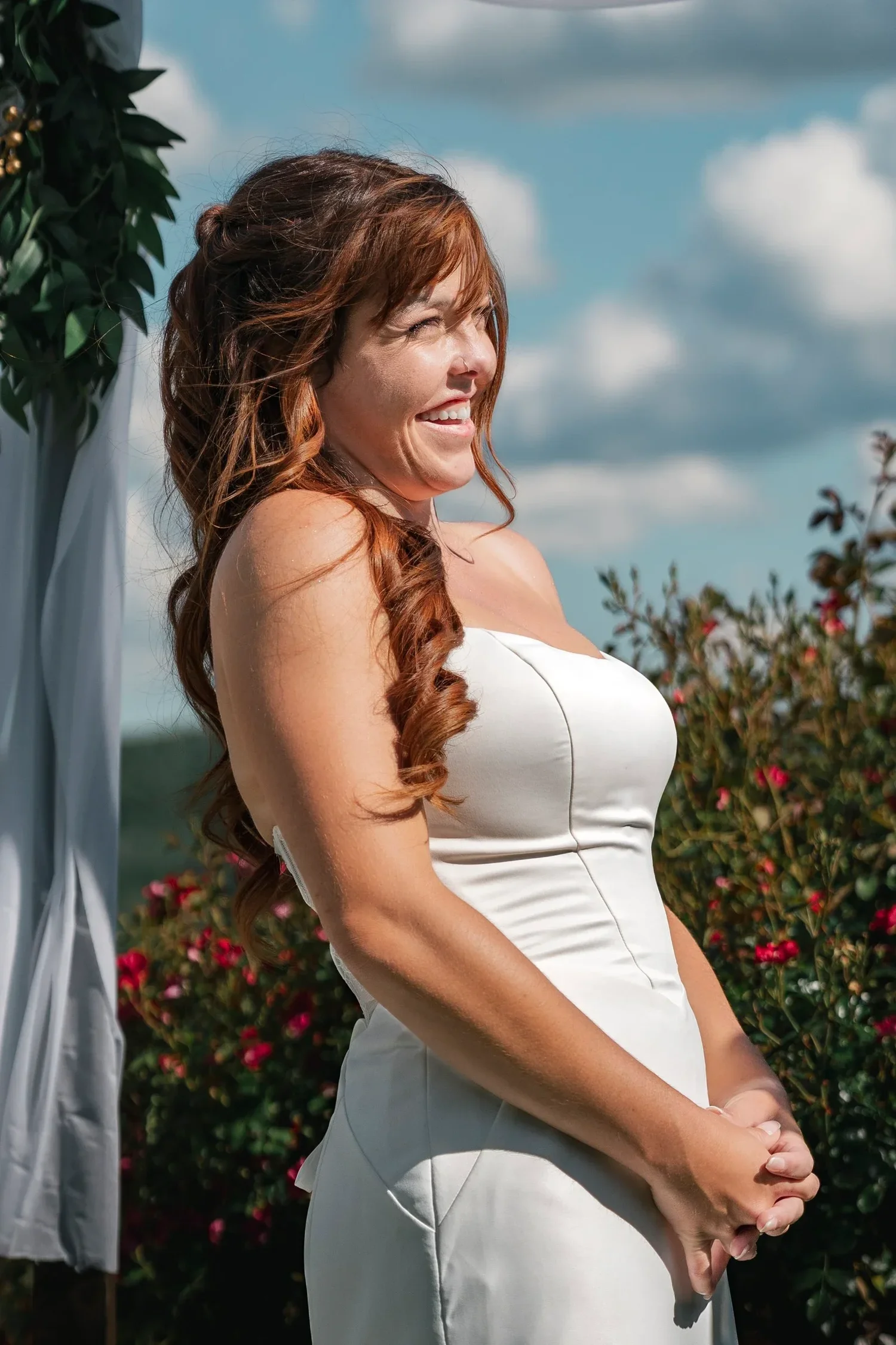 Danielle smiling during the ceremony at Traditions at the Glen, red shrubs and a cloudy blue sky behind her