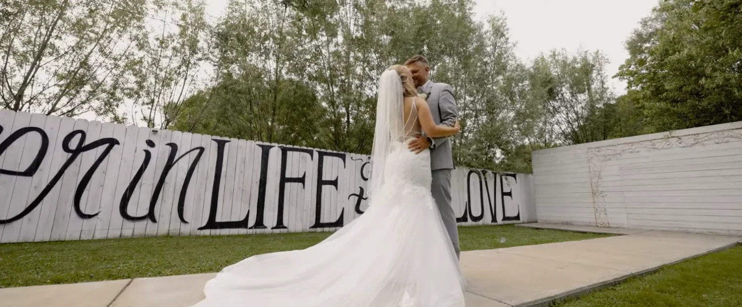 A frame from Jennifer and Alex's wedding film: the couple embracing on a lawn in front of a tall white fence painted with the words Life and Love
