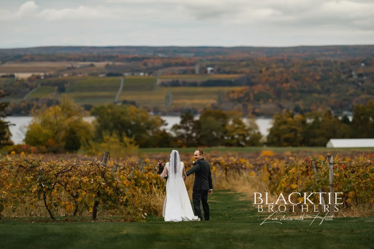 Anya and Gabe standing together in a row of golden vines at Glenora Wine Cellars, with the vineyard slopes and Seneca Lake below them