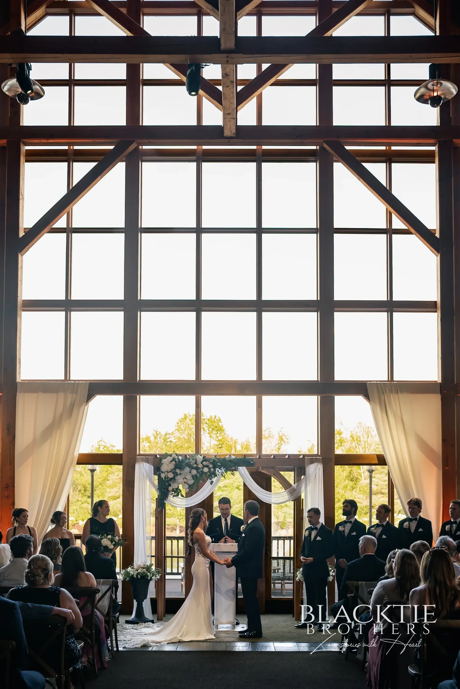 A wedding ceremony at the floor to ceiling window wall inside Allyn Lodge, guests seated under the timber frame.