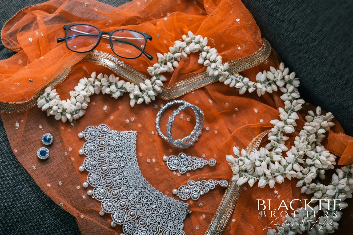 A silver choker, earrings and bangles laid out on an orange scarf with strands of white flower buds and a pair of glasses