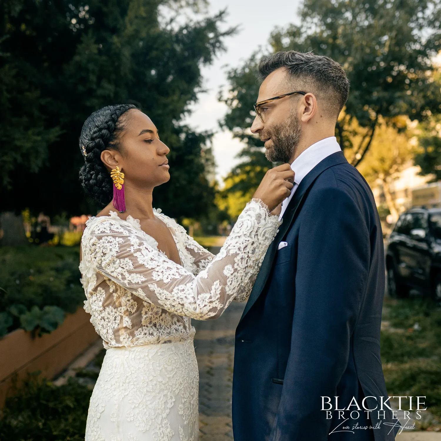 Ciara straightening Drew's bow tie on a leafy Brooklyn sidewalk, both in the warm afternoon light