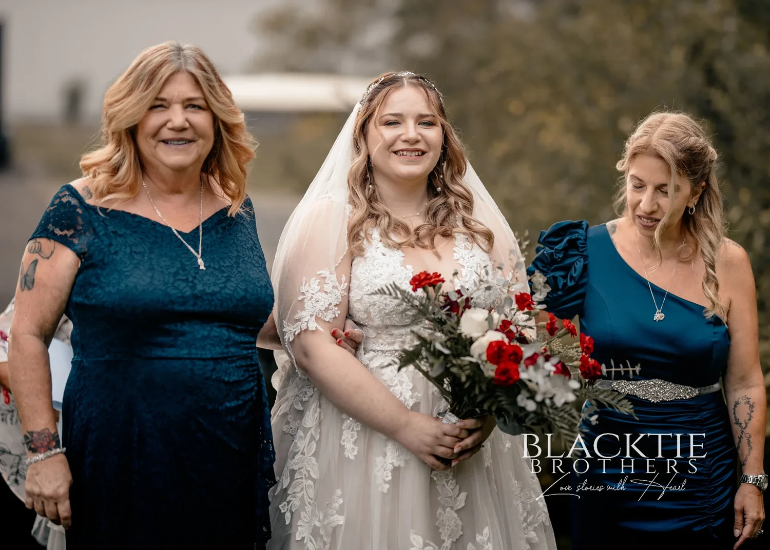 Jennifer in her veil and lace gown, smiling, walking arm in arm between two women in navy dresses, holding a red and white bouquet