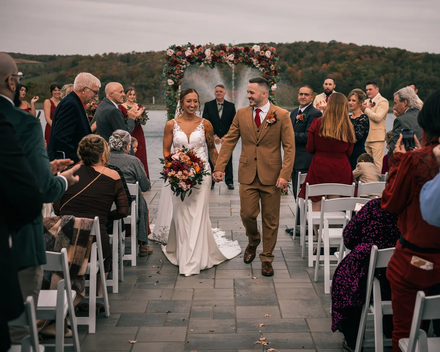 Heather and Vincent walking back up the aisle as guests cheer, the floral arch and the pond behind them