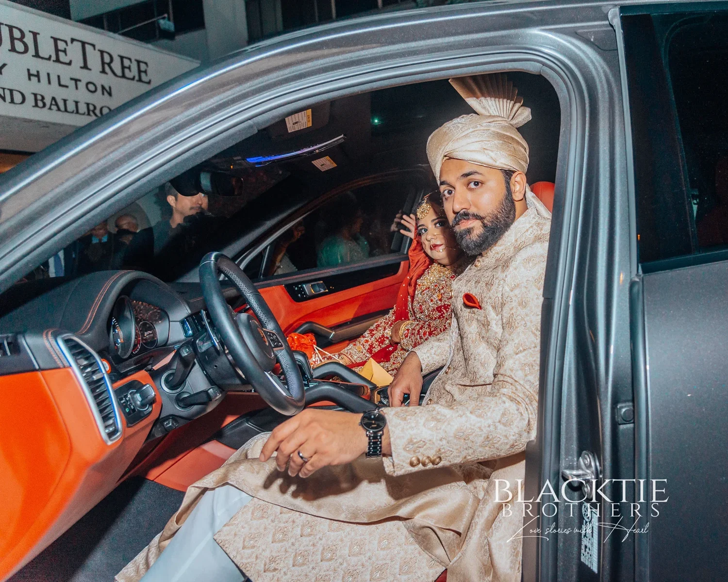 The groom smiling from the driver’s seat of a car with an orange interior, the bride beside him, and a DoubleTree by Hilton sign visible behind the car at night