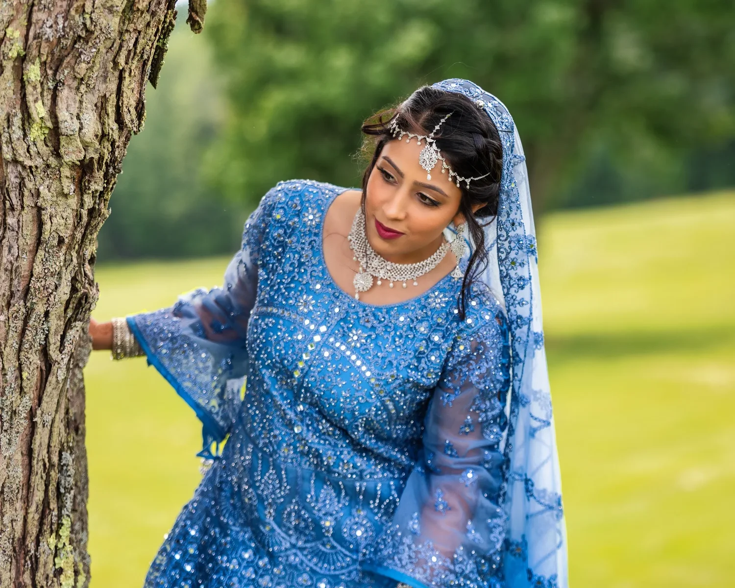 A bride in a pale blue embroidered gown and veil leaning against a tree trunk on the lawn at Traditions at the Glen