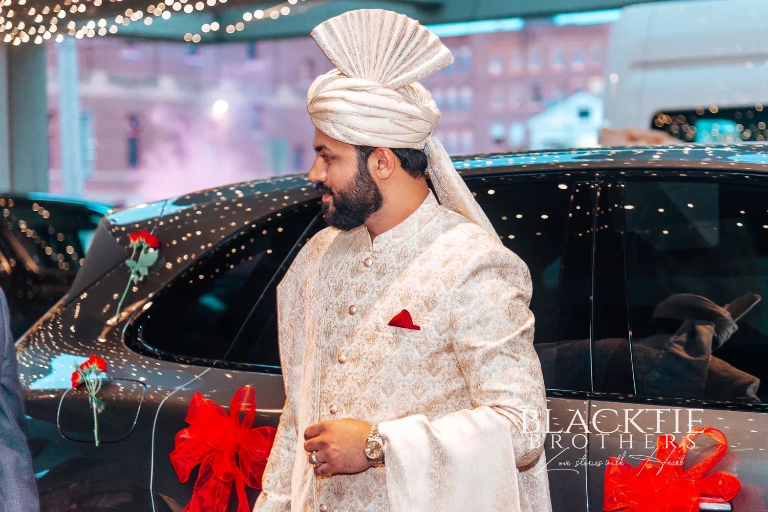 The groom in an ivory sherwani and turban stepping out of a car decorated with red ribbon under string lights at the hotel entrance