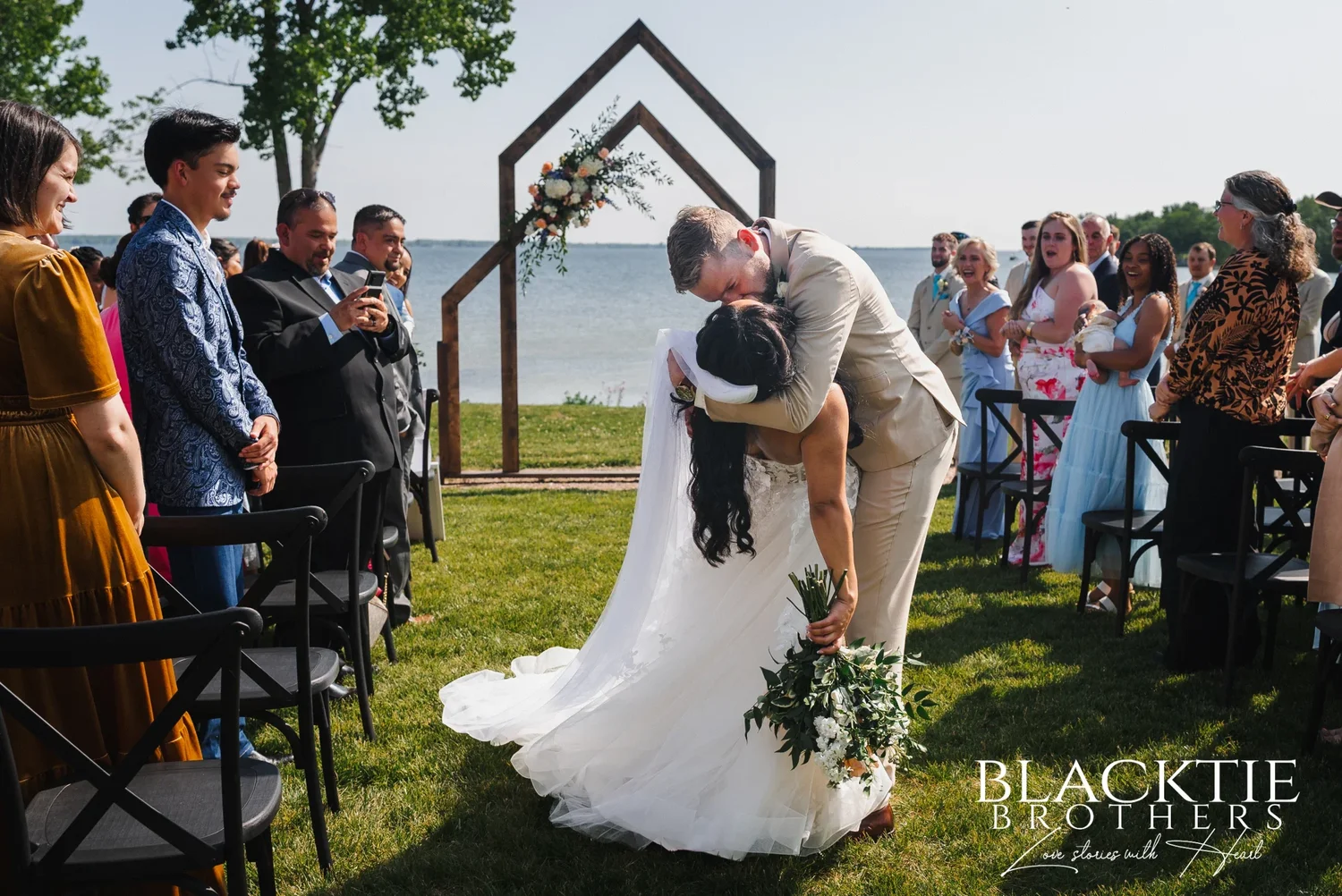 Josh dipping Carolina into a kiss in the aisle while guests stand, smile and film on their phones, the arch and the lake behind them