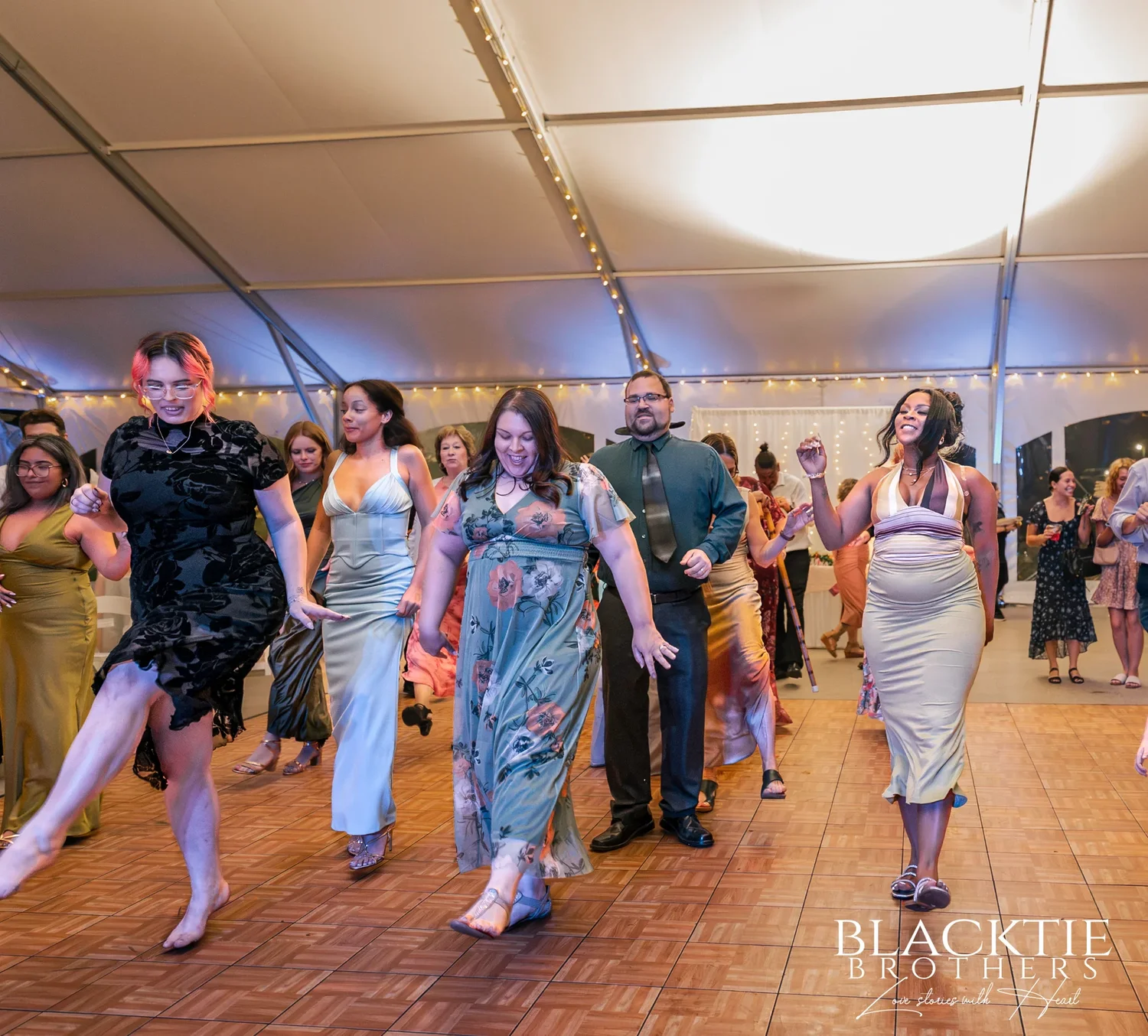 Wedding guests dancing on the wooden dance floor under the lit ceiling of the reception tent at Traditions at the Glen at night