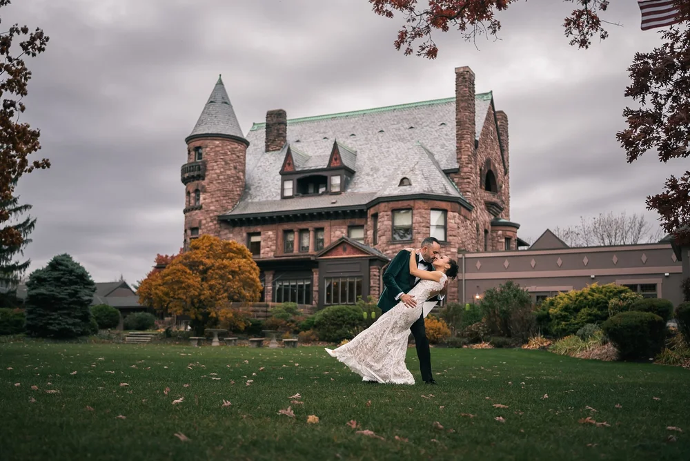 A couple kissing on the lawn in front of Belhurst Castle in Geneva, New York, its red stone turret behind them under an autumn sky