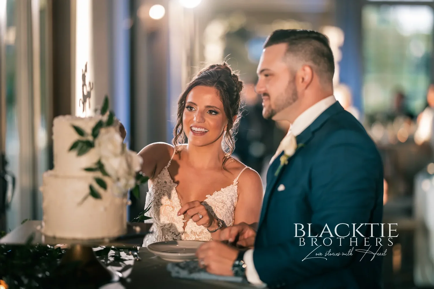 Taylor and Jake cutting their two-tier white wedding cake with greenery on it beside the tall windows, both laughing