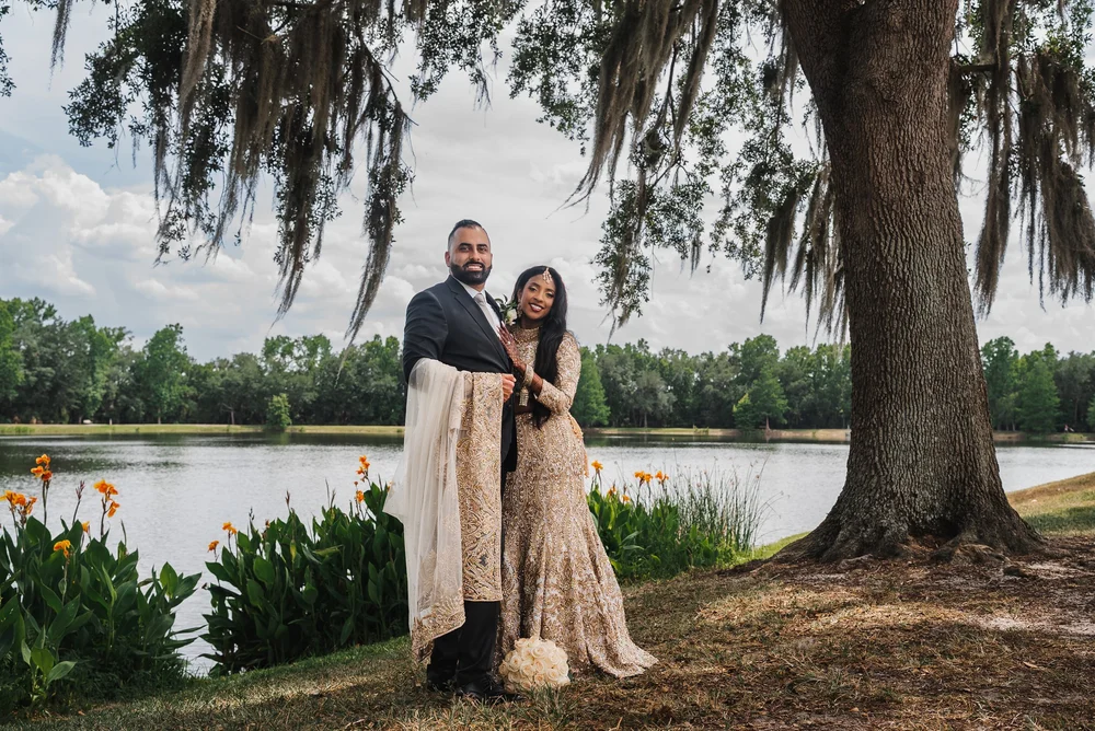 Bride and groom standing together beside a lake beneath a moss-draped tree, with flowers along the shore.