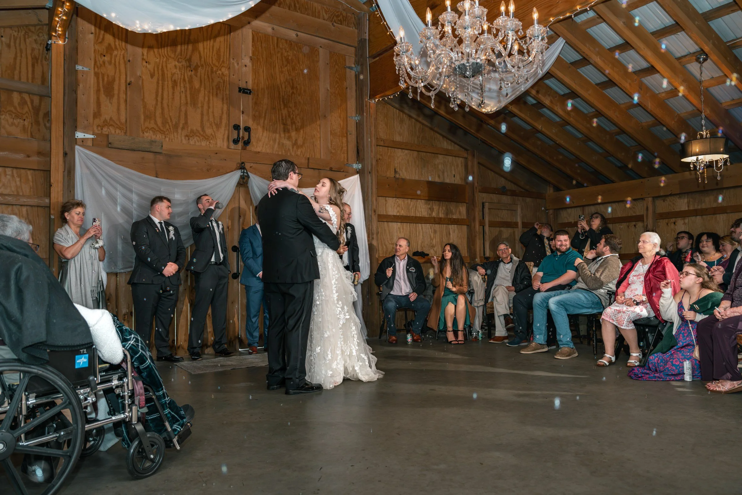 Lindsey and Noah’s first dance inside the Black Barn under the crystal chandelier, white drapes and greenery behind them and family and friends around the room