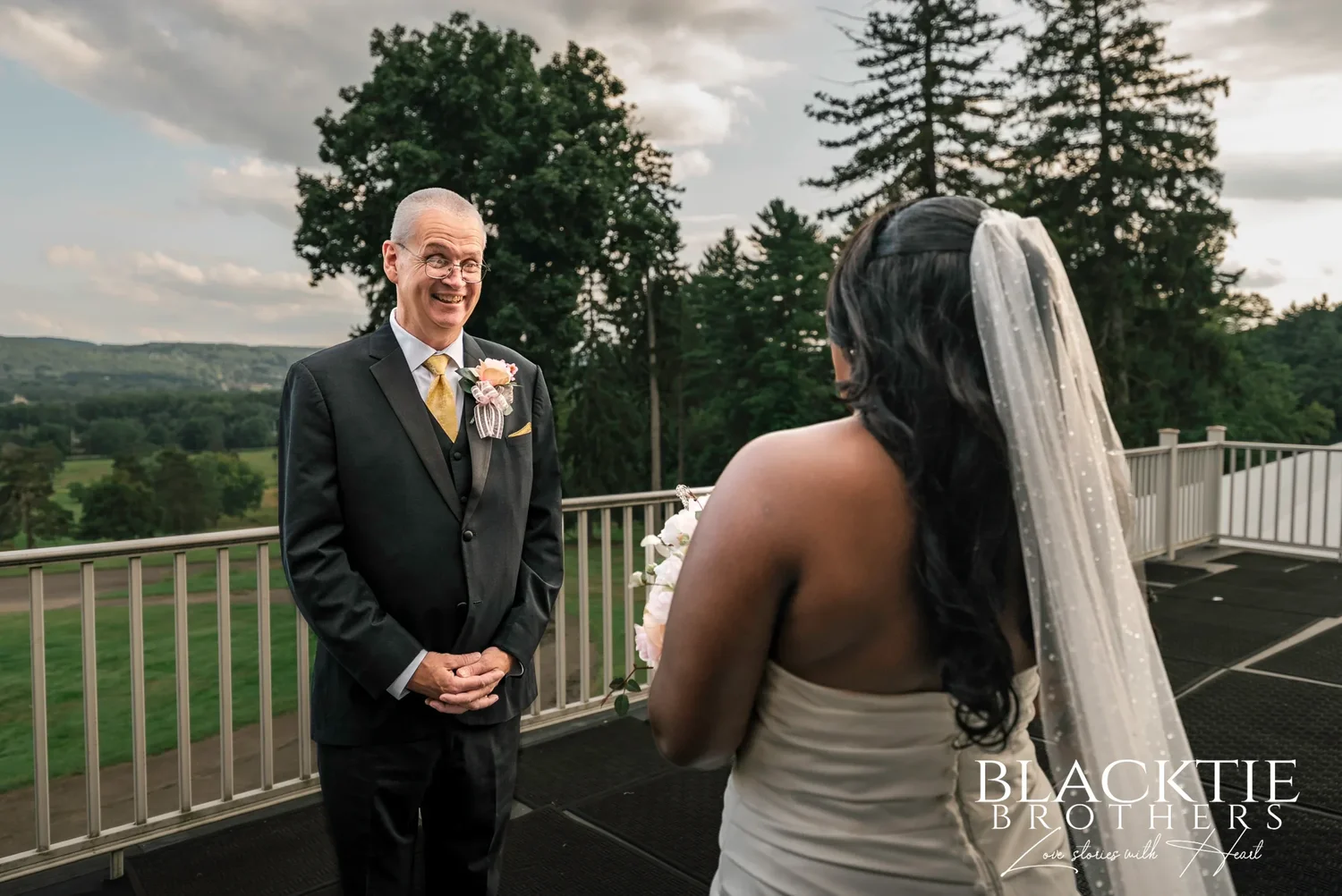 A bride in a long veil facing her father on the mansion balcony at Traditions at the Glen for their first look, with the valley and tree line behind him
