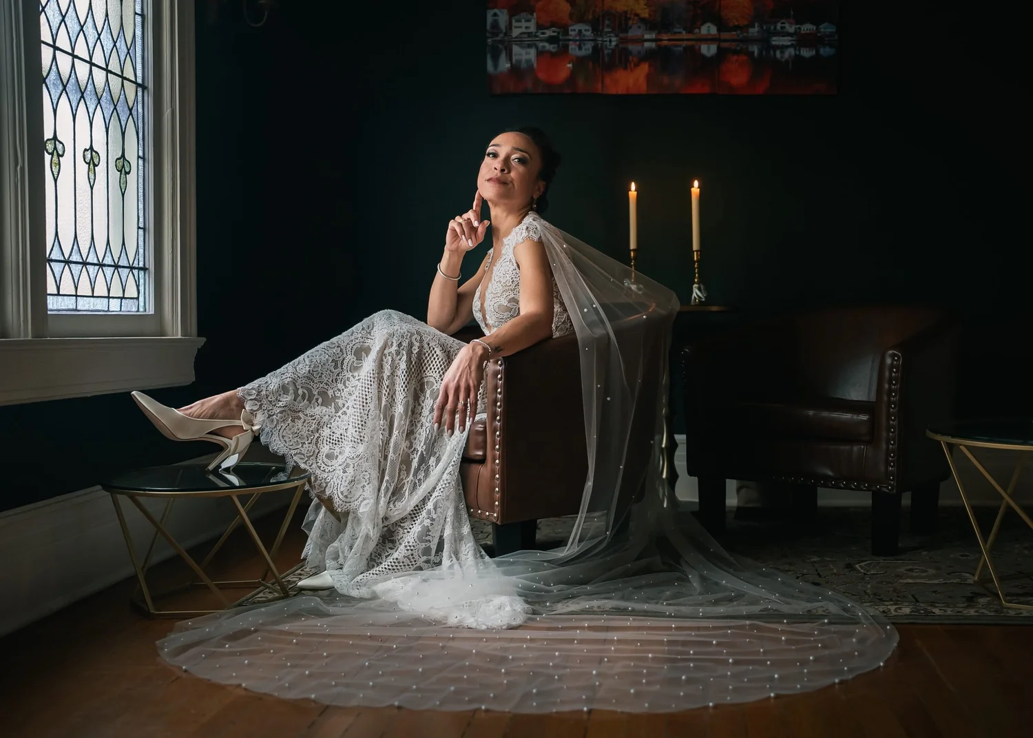 Shannon lounging sideways in a leather armchair by candlelight, finger at her chin and veil pooled on the floor, at the rental house where she got ready