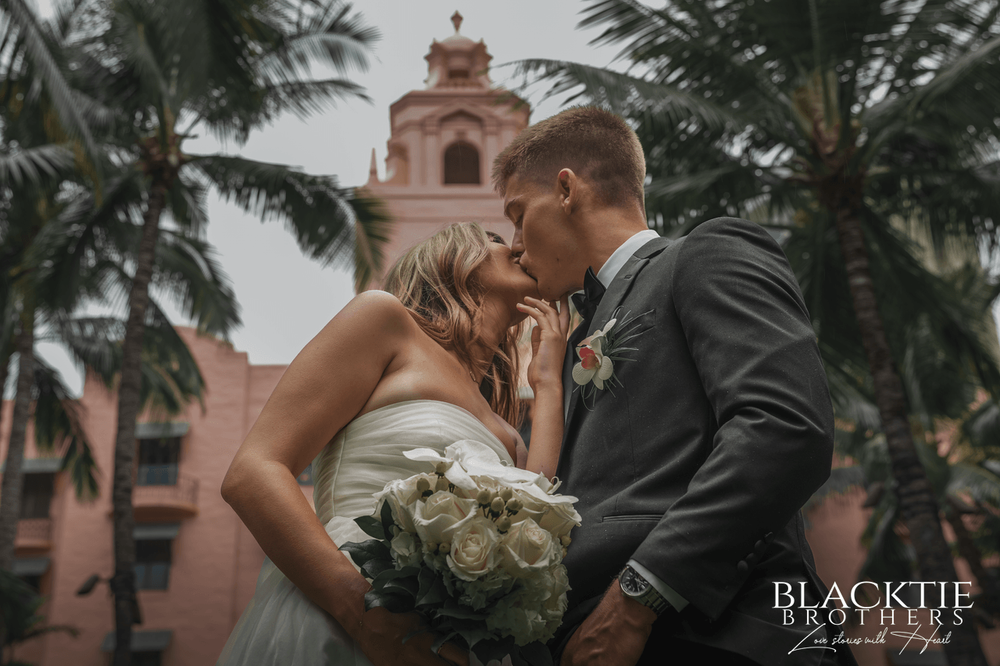 Bride and groom kissing with a bouquet beneath palms and pink architecture.