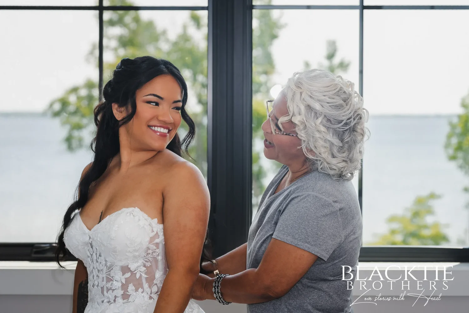 Carolina smiling over her shoulder while a woman with silver hair fastens the back of her strapless lace dress beside a window overlooking the water