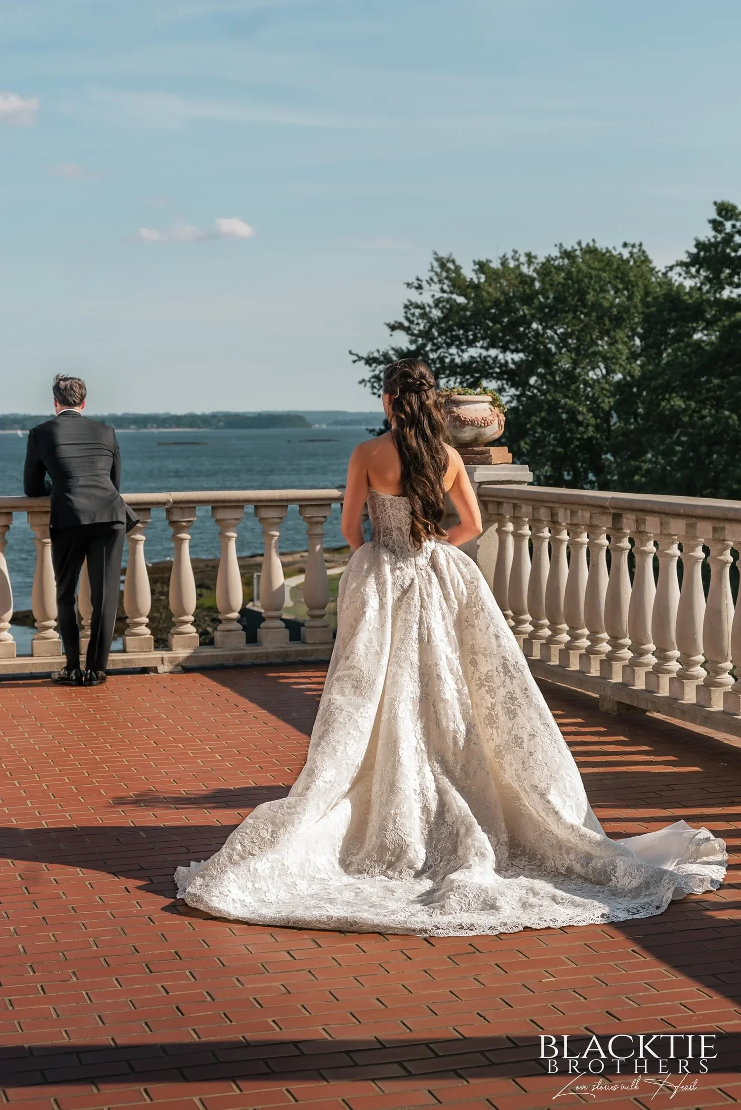 Leah in her strapless lace gown and long train walking across the brick terrace toward Nicholas, who stands at the white balustrade with his back turned, looking out over the Sound