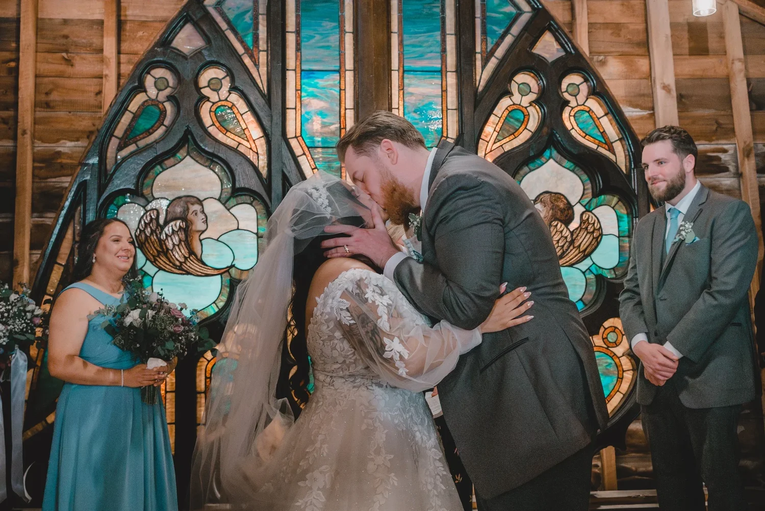 Jessica and Christian kissing in front of the stained glass window in the Loft Chapel while members of the wedding party look on