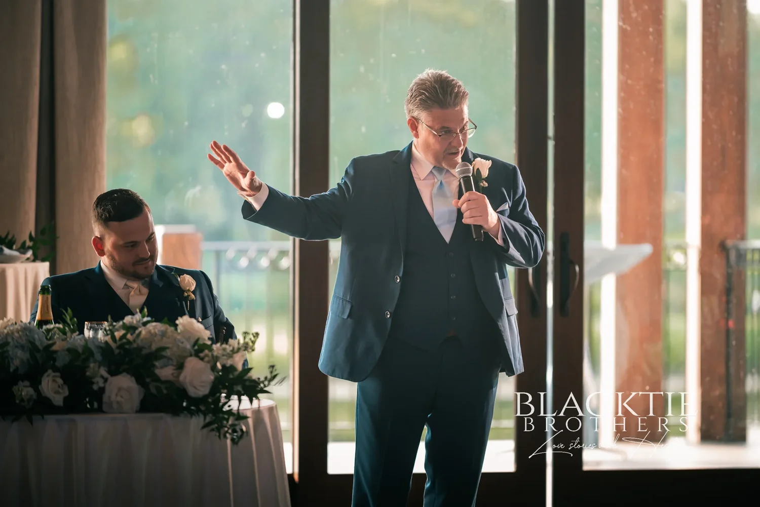 A father giving a toast with a microphone beside the sweetheart table, the tall windows and the course behind him