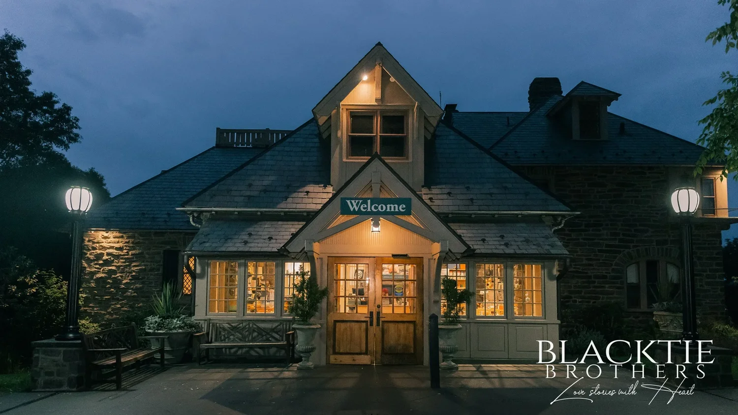 The stone carriage house at Morris Arboretum at dusk, lamps glowing on either side of the wooden doors under a Welcome sign