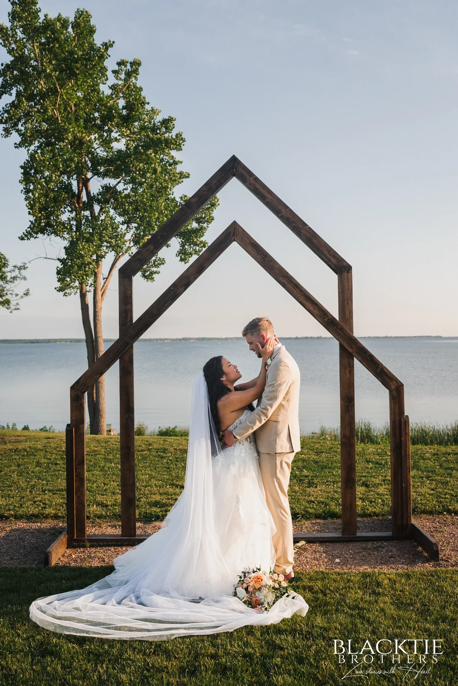 Carolina and Josh holding each other under the wooden arch at the shoreline in evening light, her veil and train spread across the grass, the lake behind them