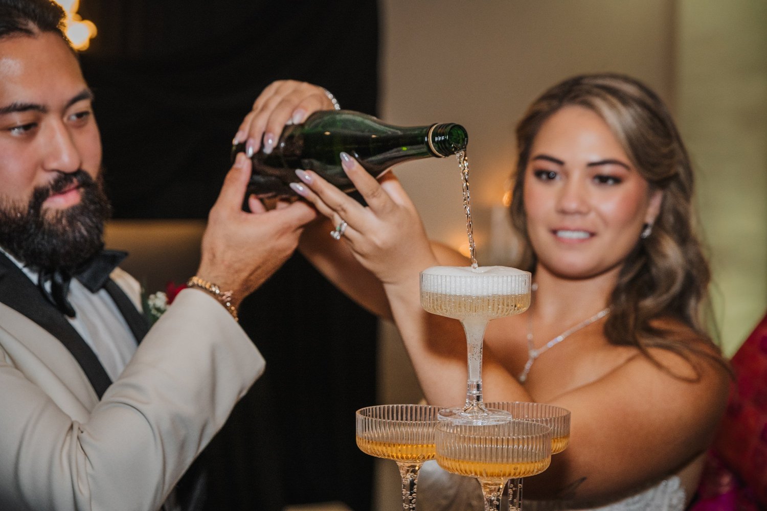 Alice pouring champagne into the top glass of a champagne tower while Mitchell holds the bottle with her