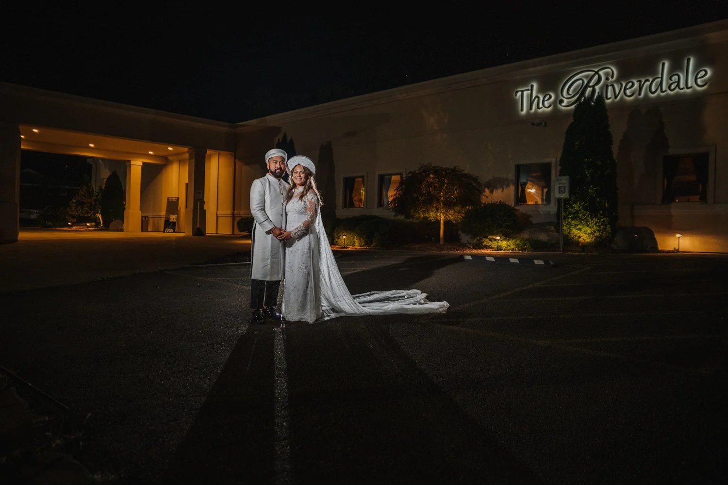 Alice and Mitchell outside The Riverdale at night, the entrance portico glowing and the lit Riverdale sign on the wall beside them