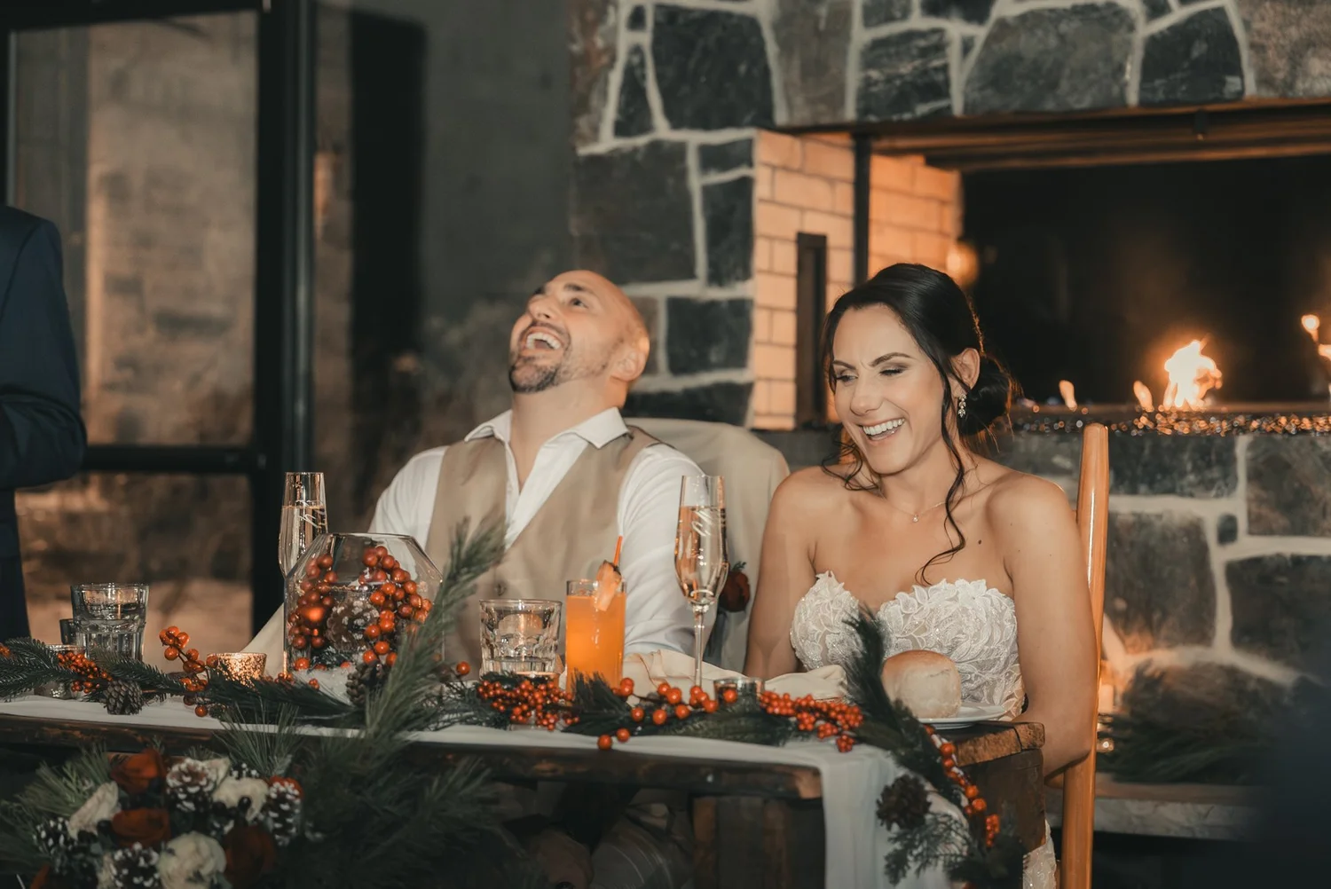 Jill and Nick laughing at their Christmas wedding sweetheart table, decorated with holiday greenery beside a stone fireplace at The Sinclair of Skaneateles