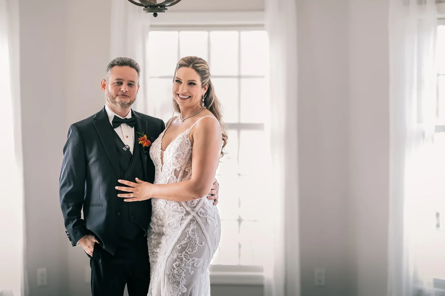 Charissa and Andrew standing together in soft window light inside the Manor House, sheer curtains on either side