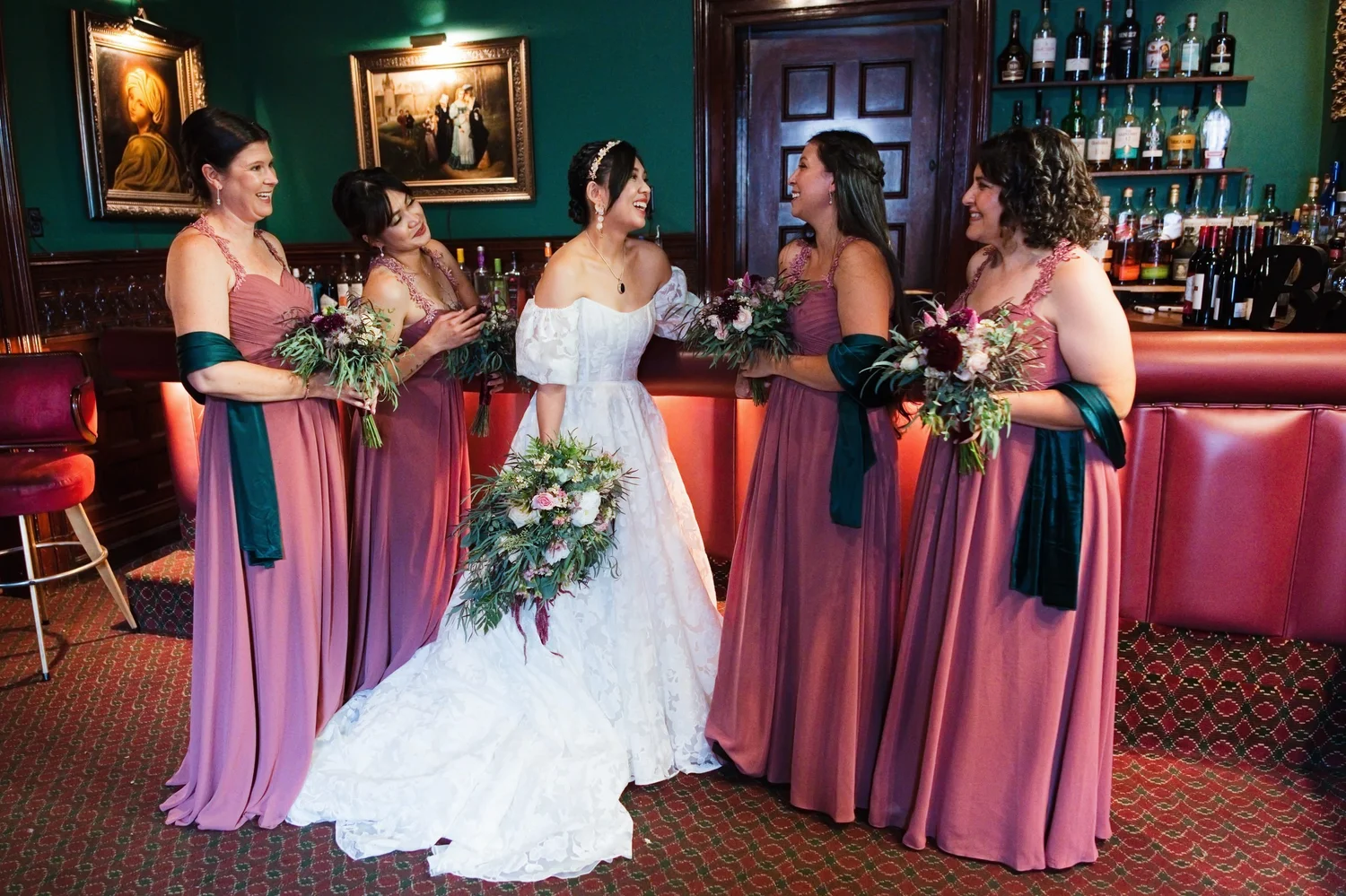 Marseille laughing with four bridesmaids in rose and mauve dresses and green wraps at the bar, teal walls and red leather behind them