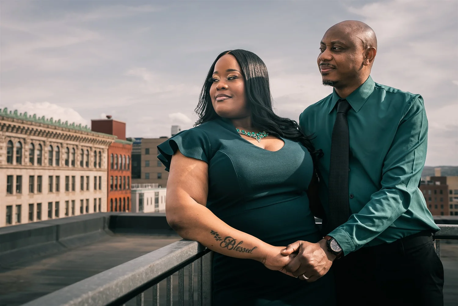 Tiffany and David in coordinated green outfits stand at a terrace railing with downtown Syracuse behind them during their second engagement session.