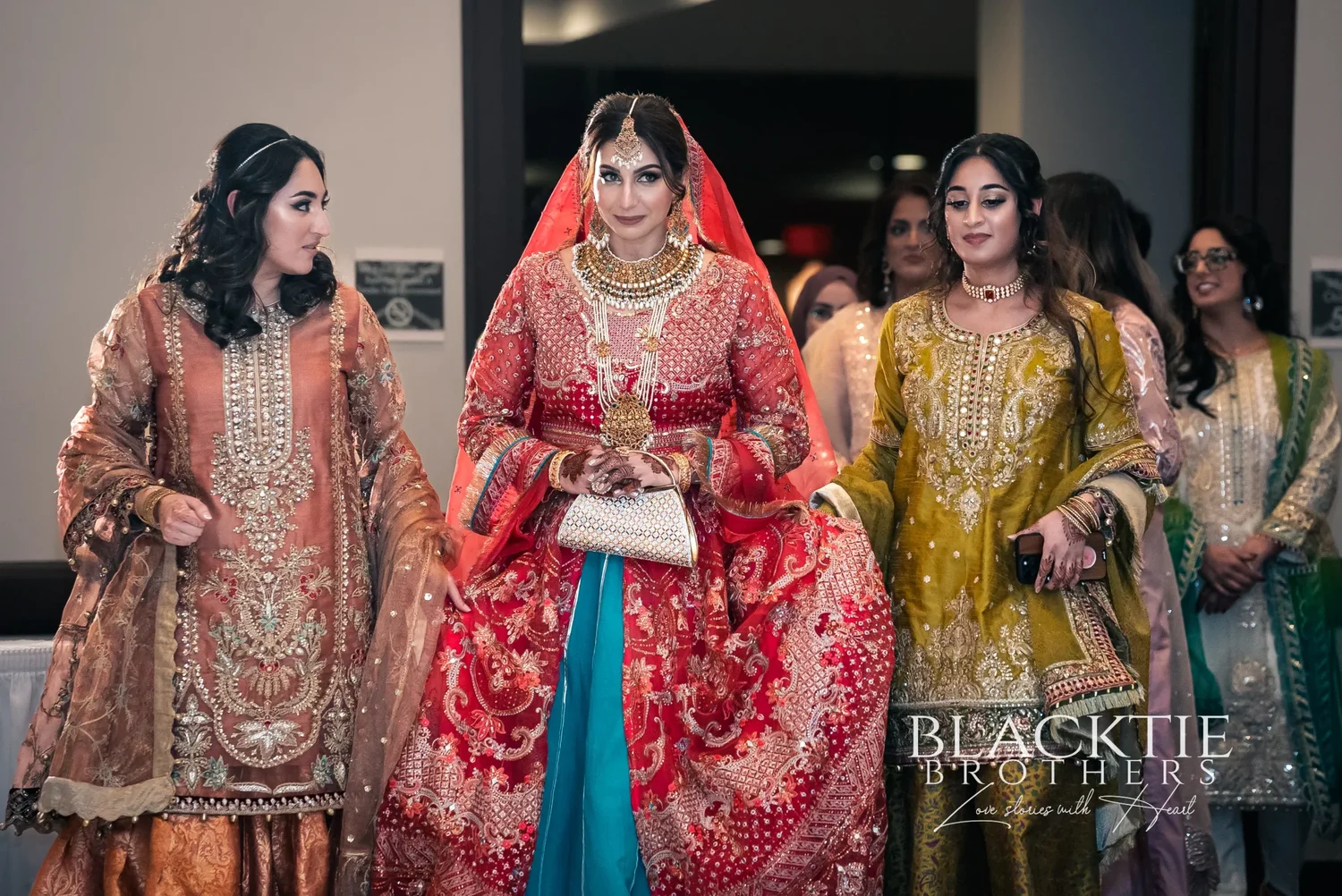 Jia walking in to her Nikkah between two women in embroidered peach and mustard outfits, all three smiling, in a corridor at the hotel