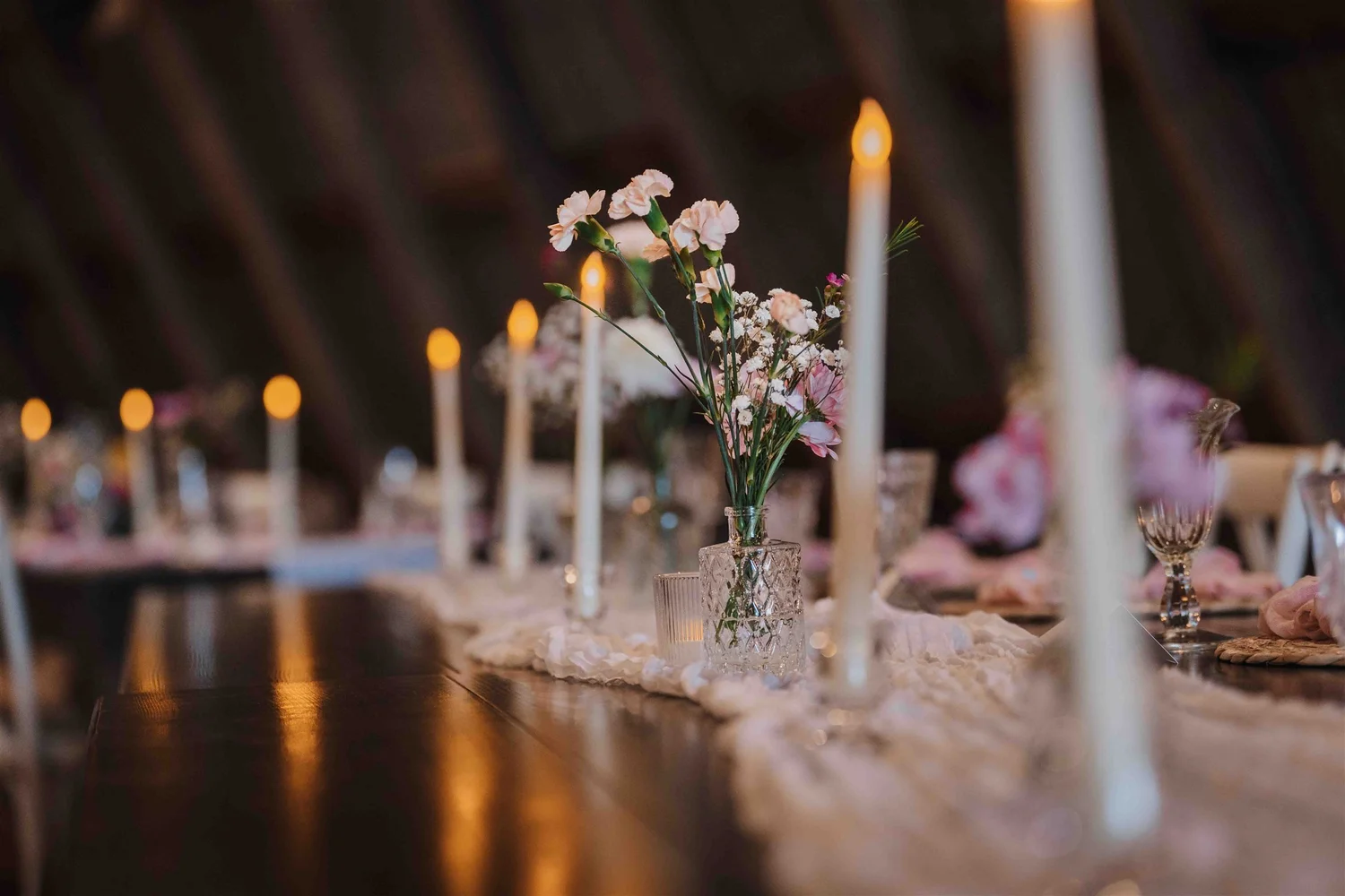 Reception table set with white candles, small pink flower arrangements and a pale fabric runner.