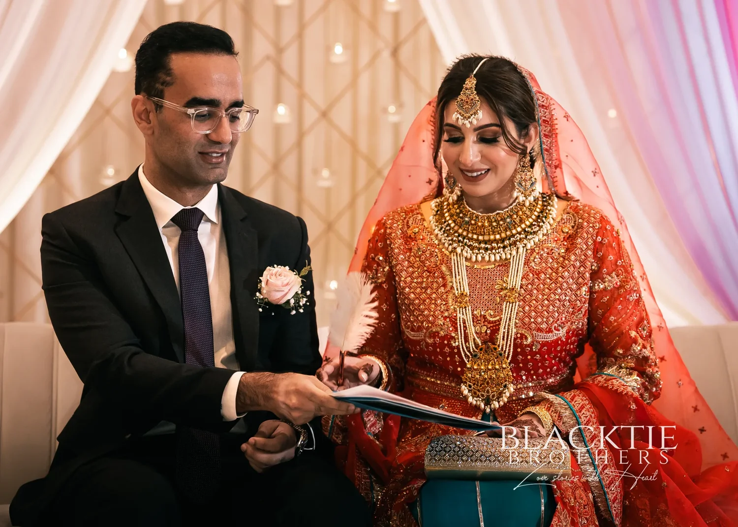 Rizwan signing the Nikkah papers with a white feather pen while Jia, seated beside him in red and gold, looks on smiling