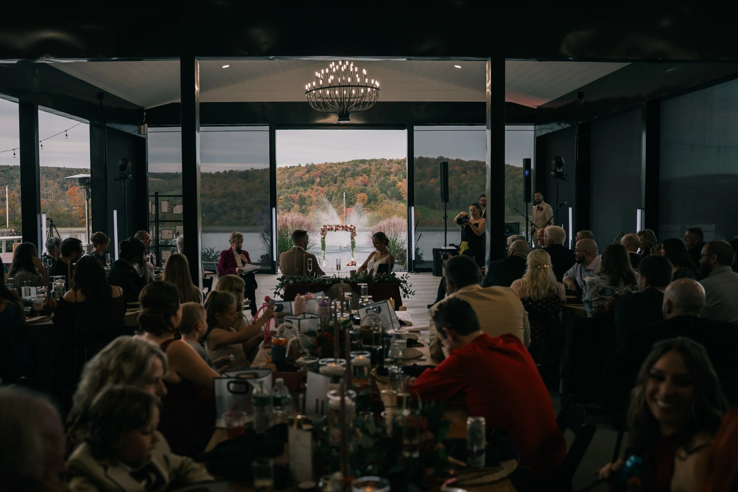 The glass-walled pavilion during the reception, guests at their tables under the chandelier, with the pond and the hills of the Endless Mountains through the windows