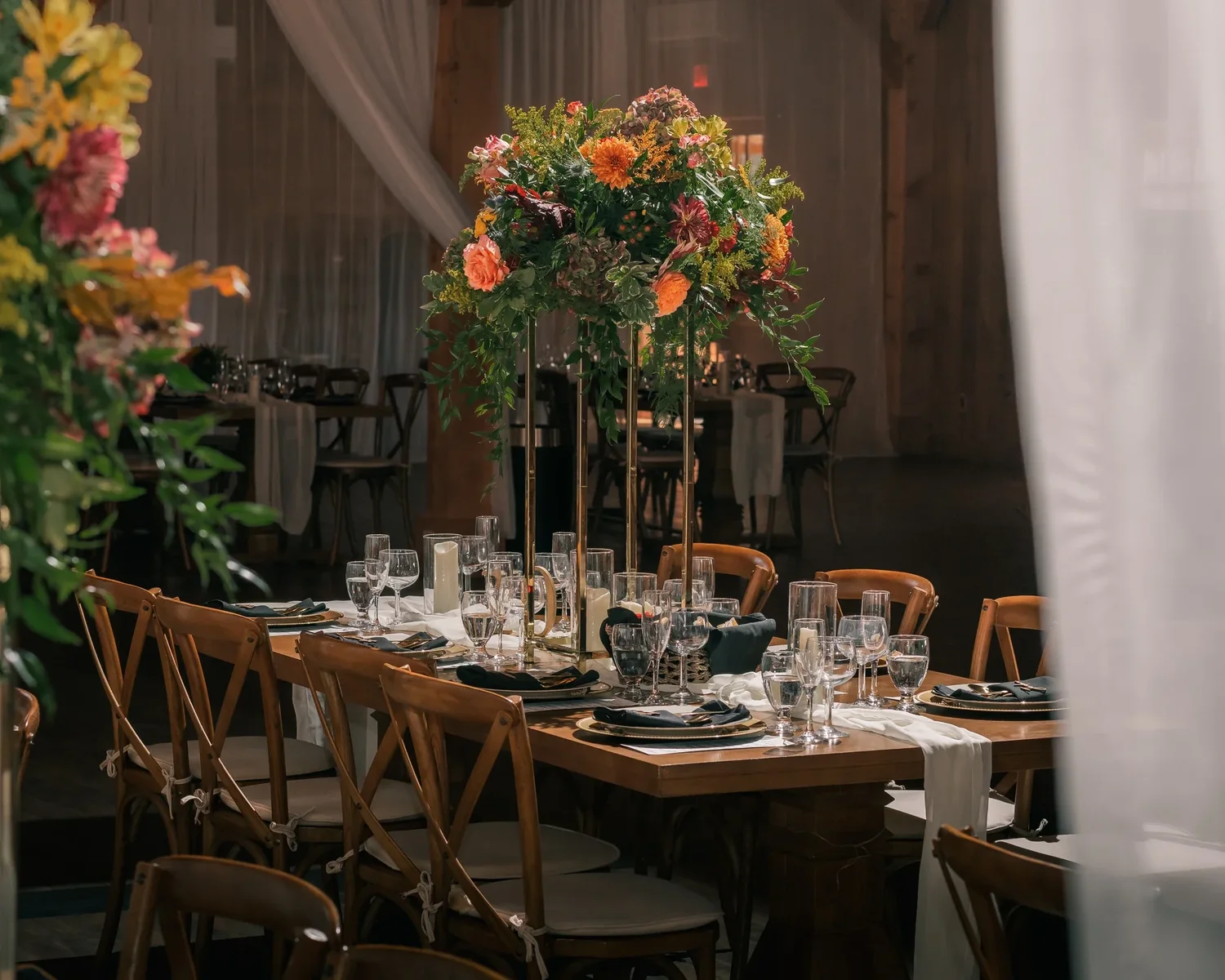 A long farm table in the Barn set for dinner with gold chargers, glassware and tall arrangements of orange and green autumn flowers